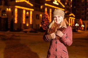 Street portrait of smiling beautiful young woman on the festive Christmas fair. Lady wearing classic stylish winter knitted clothes. Model looking at camera. Magic snowfall effect. Close up photo