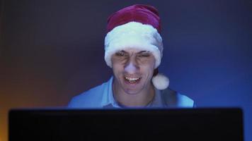 Happy man in santa claus hat smiles and communicates on a video call with friends using a laptop. The room has neon lighting. Online leisure concept.