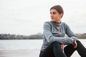 Young sportive girl sitting on the ground outdoors near lake at daytime photo