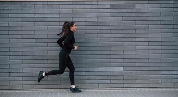 Young sportive girl in black sportswear running outdoors near gray wall photo