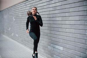 joven deportista con ropa deportiva negra corriendo al aire libre cerca de la pared gris foto