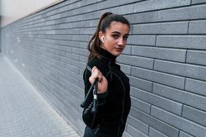 Young sportive girl in black sportswear standing with jump rope in hands outdoors near gray wall photo