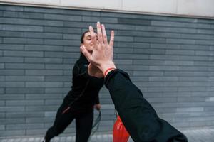 Young sportive girl in black sportswear standing with jump rope in hands outdoors near gray wall and giving high five photo