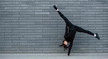 Young sportive girl in black sportswear doing hard handstand exercises outdoors near gray wall photo