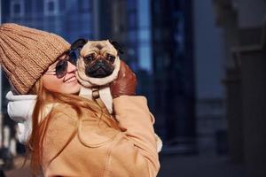 Woman in warm clothes have holding her little pug dog on hands near business building that on background photo