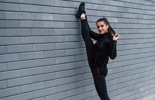 joven deportiva en ropa deportiva negra haciendo piernas estiradas al aire libre usando una pared gris foto