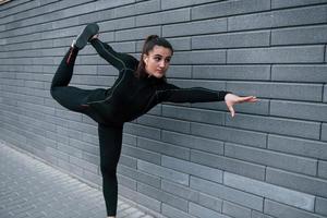 Young sportive girl in black sportswear outdoors doing yoga exercises near gray wall photo