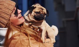 Woman in warm clothes have holding her little pug dog on hands near business building that on background photo