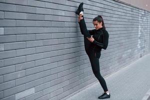 Young sportive girl with phone in black sportswear doing legs stretching outdoors by using gray wall photo