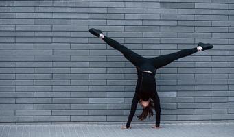 Young sportive girl in black sportswear doing hard handstand exercises outdoors near gray wall photo