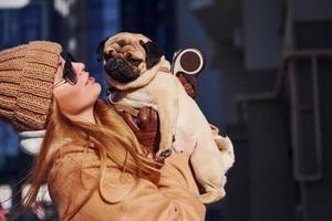 Woman in warm clothes have holding her little pug dog on hands near business building that on background photo