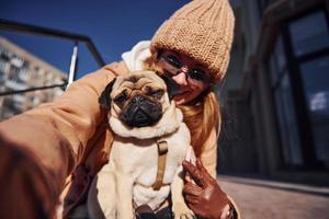 Woman in warm clothes make selfie with her little pug dog near business building that on background photo