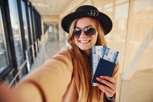 joven pasajera con ropa abrigada que muestra boletos en la sala del aeropuerto foto