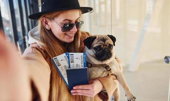 Young female passenger in warm clothes holding tickets and cute dog in hands in airport hall photo