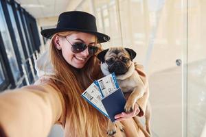 Young female passenger in warm clothes holding tickets and cute dog in hands in airport hall photo