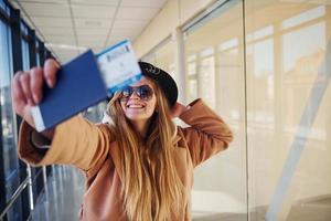 joven pasajera con ropa abrigada que muestra boletos en la sala del aeropuerto foto