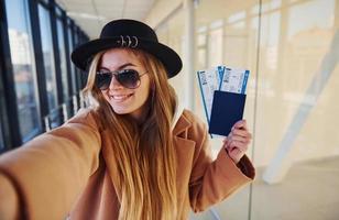 Young female passenger in warm clothes showing tickets in airport hall photo