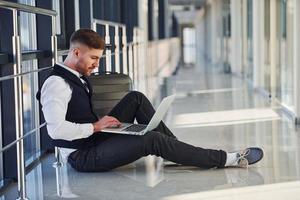 Young male passenger in elegant formal clothes sitting with laptop in the airport hall photo