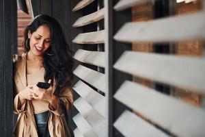 Woman with black curly hair standing near wooden windows with phone in hands photo