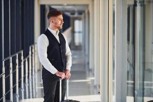 Young male passenger in elegant formal clothes is in the airport hall standing with baggage photo