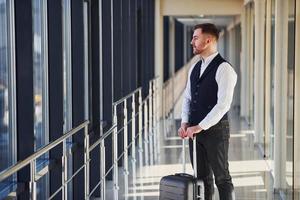 un joven pasajero con ropa elegante y formal está en la sala del aeropuerto de pie con equipaje foto