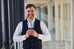 joven pasajero con ropa elegante y formal sonriendo y sosteniendo boletos en las manos en la sala del aeropuerto foto