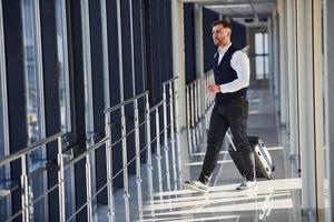 Young male passenger in elegant formal clothes is in the airport hall walking with baggage photo