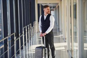 un joven pasajero con ropa elegante y formal está en la sala del aeropuerto de pie con equipaje foto