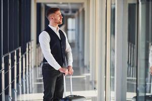 Young male passenger in elegant formal clothes is in the airport hall standing with baggage photo