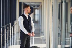 un joven pasajero con ropa elegante y formal está en la sala del aeropuerto de pie con equipaje foto