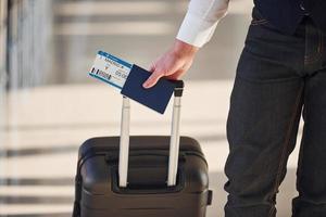 vista de cerca del pasajero masculino con ropa elegante y formal que está en la sala del aeropuerto con equipaje y boletos foto