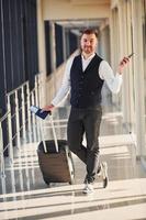 Male passenger in elegant formal clothes is in the airport hall with baggage, tickets and phone photo