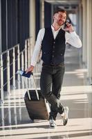 el pasajero masculino con ropa formal elegante está en la sala del aeropuerto con equipaje, boletos y teléfono foto