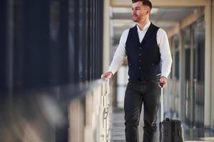 Young male passenger in elegant formal clothes is in the airport hall walking with baggage photo