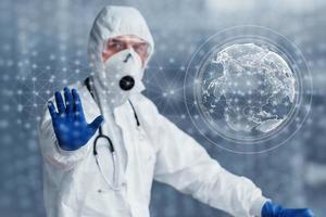 Digital world map on picture. Male doctor scientist in lab coat, defensive eyewear and mask standing indoors photo