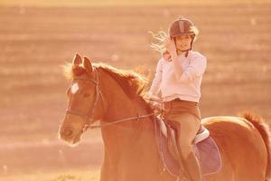 Young woman in protective hat riding her horse in agriculture field at sunny daytime photo
