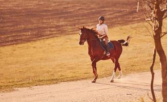 mujer joven con sombrero protector montando su caballo en el campo agrícola durante el día soleado foto