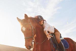 Young woman in protective hat with her horse in agriculture field at sunny daytime photo