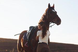 Beautiful sunshine. Young woman standing with her horse in agriculture field at daytime photo