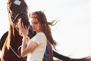 Beautiful sunshine. Young woman standing with her horse in agriculture field at daytime photo