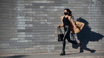 In protective mask. Beautiful brunette with curly hair and in black clothes running outdoors near wall photo