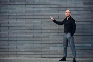 hombre elegante de moda parado al aire libre contra la pared gris y muestra a la izquierda con el dedo foto