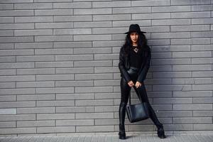 hermosa morena con cabello rizado y ropa negra sosteniendo una bolsa al aire libre cerca de la pared foto