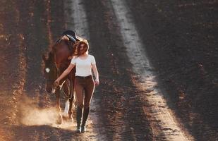 Young woman in protective hat walking with her horse in agriculture field at sunny daytime photo