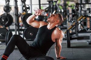 tomando un descanso y bebiendo agua. joven deportista fuerte vestido de negro tiene un día de entrenamiento en el gimnasio foto