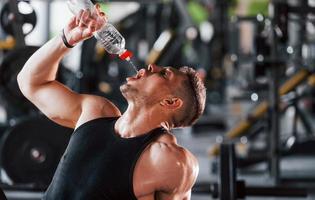 tomando un descanso y bebiendo agua. joven deportista fuerte vestido de negro tiene un día de entrenamiento en el gimnasio foto