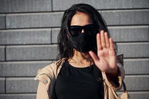 Showing stop gesture by hand. In protective mask. Beautiful brunette with curly hair and in black clothes outdoors near wall photo