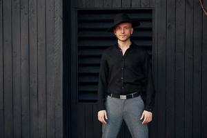 hombre con sombrero negro parado contra el exterior del edificio de madera negra foto