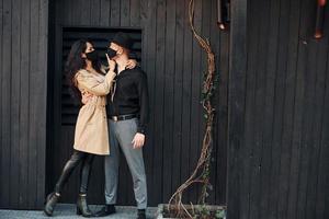 Woman with black curly hair standing against black wooden building exterior with her man in hat photo