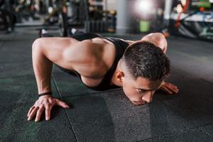 Doing push-ups. Young sportive strong man in black wear have workout day in gym photo